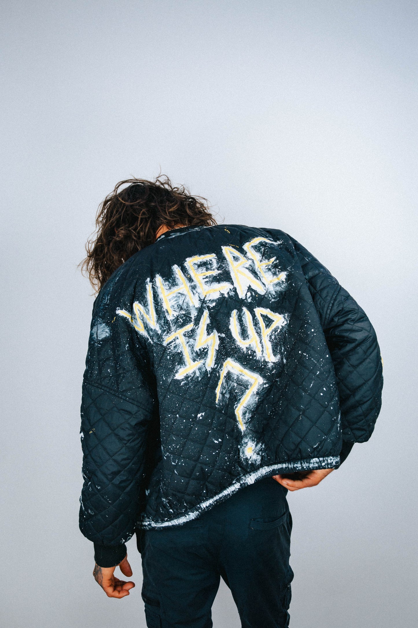 Person wearing a black bomber jacket with 'WHERE IS UP?' spray painted and hand painted text on the back against a plain background