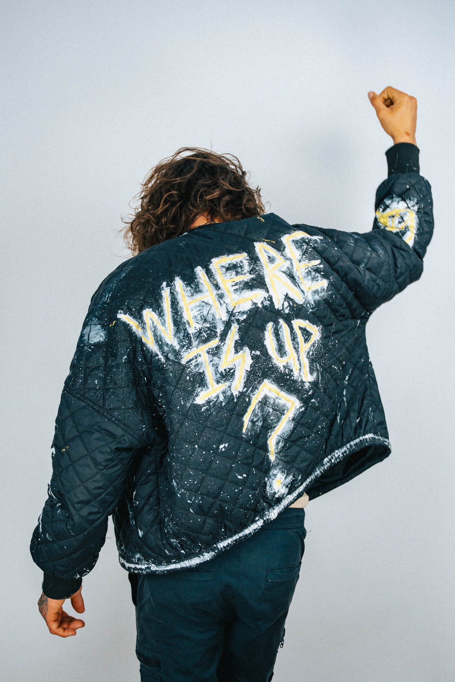 Person wearing a black quilted bomber jacket with 'WHERE IS UP?' hand painted and spray painted text on the back against a light background