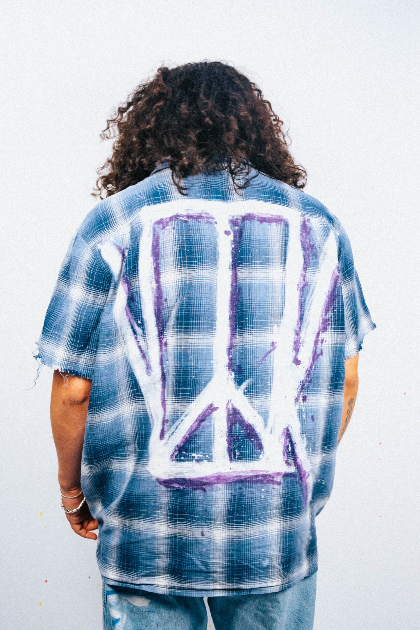 Person wearing a blue plaid shirt with a peace symbol spray painted white and purple design on the back against a white background