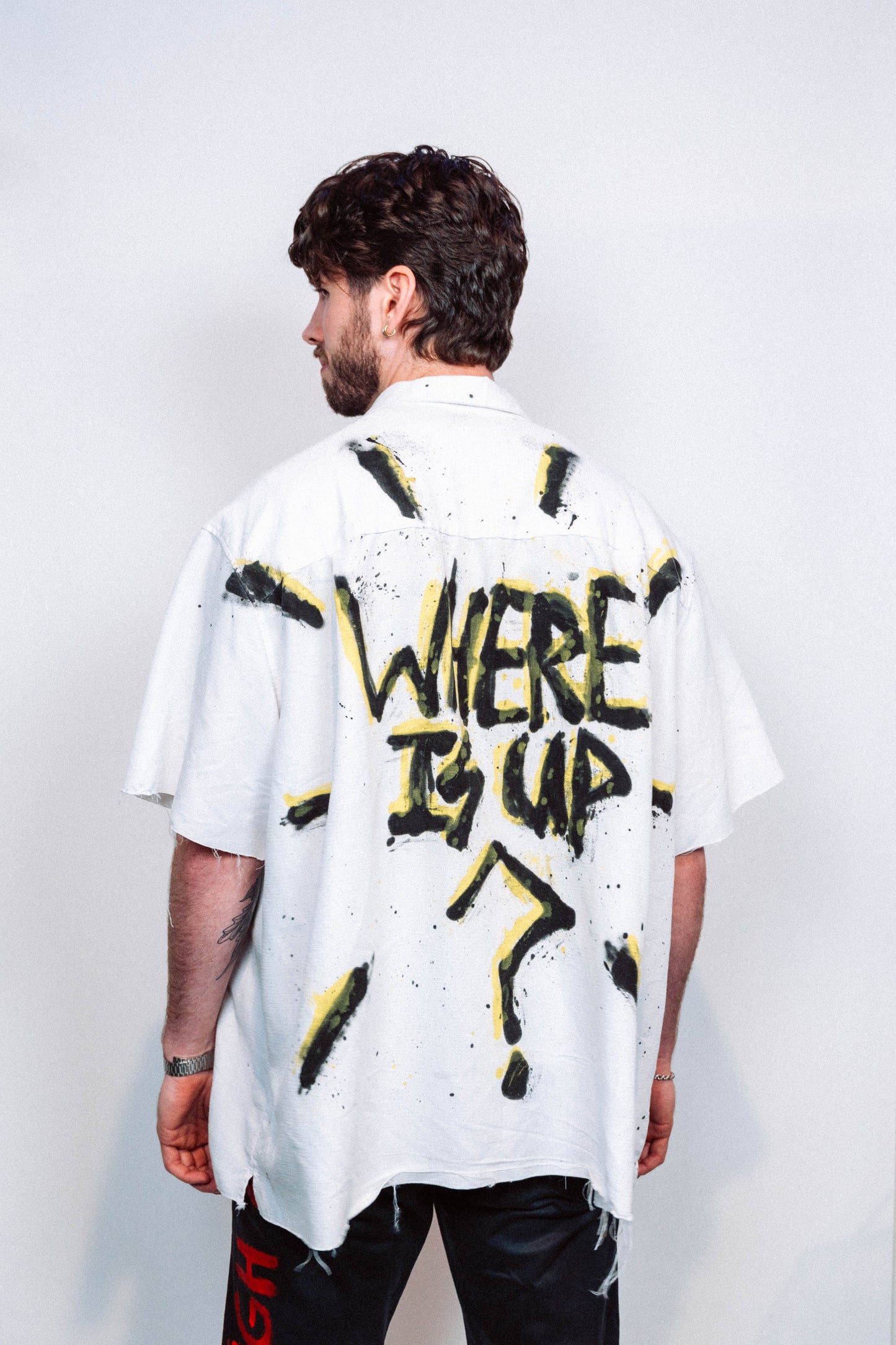 Man wearing a 1 of 1 white button up shirt with colorful spray painted designs and 1 of 1 wearable art multicolored pants against a plain background