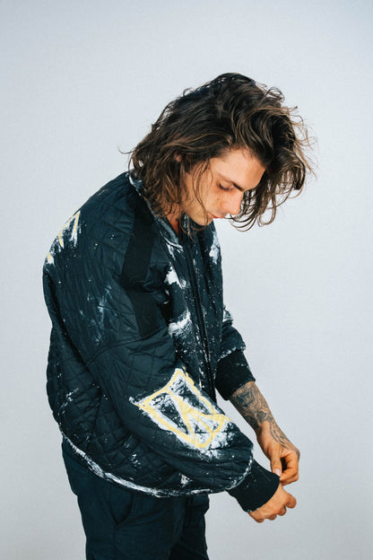 Person wearing a black quilted bomber jacket with white and yellow spray painted and hand painted designs on a plain background