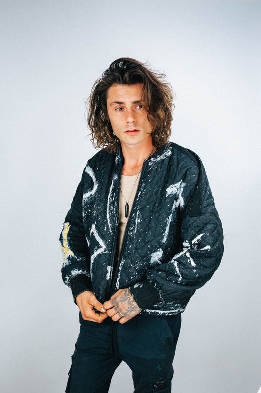 Person wearing a black bomber jacket with white and yellow spray painted designs on a plain background