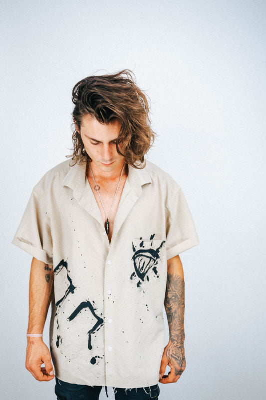 Person wearing a tan button-up shirt with black spray painted designs on a plain background