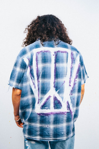 Person wearing a blue plaid shirt with a peace symbol spray painted white and purple design on the back against a white background