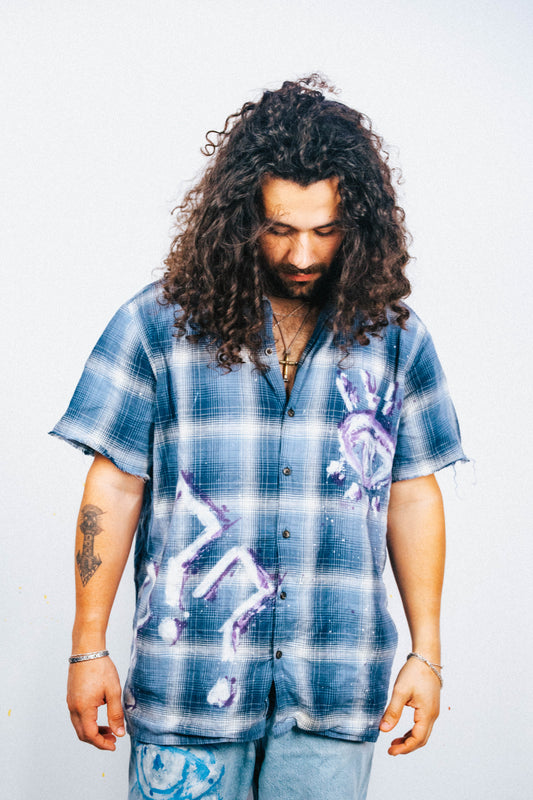 Man wearing a blue plaid shirt with white and purple spray painted graphic designs on a plain background