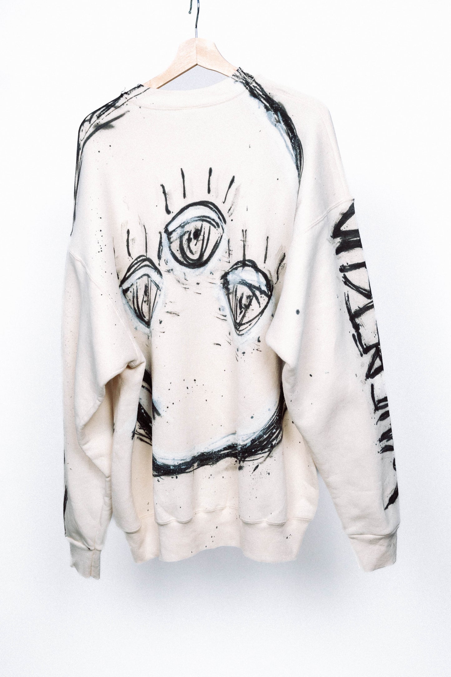 Cream colored 1 of 1 sweater with black abstract spray painted designs on a hanger against a white background