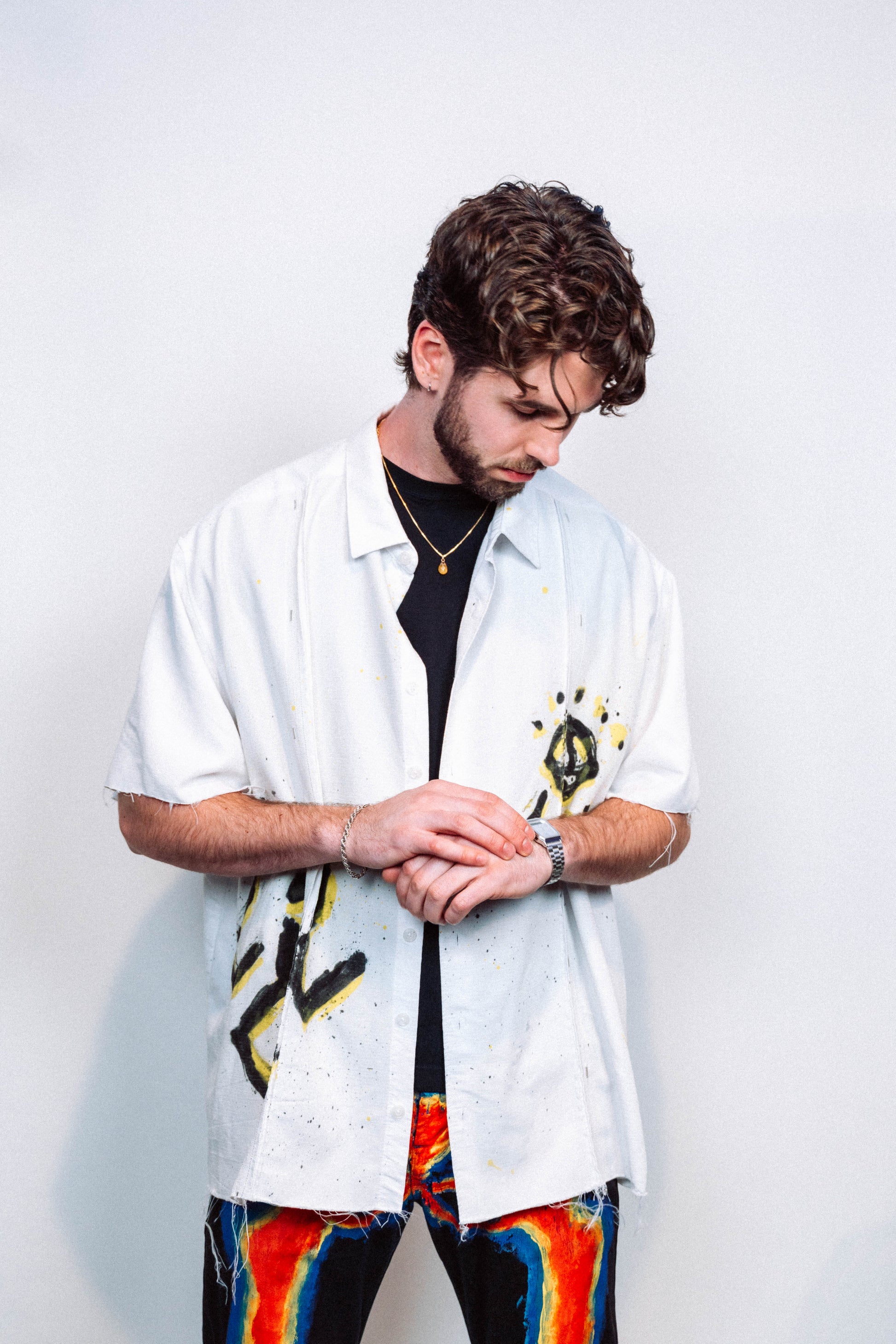 Man wearing a 1 of 1 white button up shirt with colorful spray painted designs and 1 of 1 wearable art multicolored pants against a plain background 