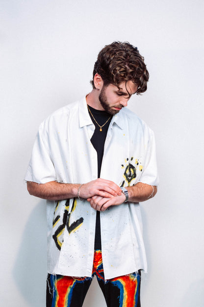 Man wearing a 1 of 1 white button up shirt with colorful spray painted designs and 1 of 1 wearable art multicolored pants against a plain background 